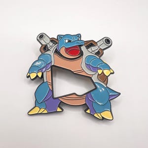 May include: A blue and brown enamel pin featuring a cartoon character with a shell on its back and two cannons on its shoulders.