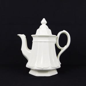 May include: A white ceramic teapot featuring a faceted, octagonal body with vertical fluted panels, a curved handle, a pointed spout, and a matching removable lid with a finial top, presented against a solid black background.