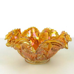 Puede incluir: Un cuenco de cristal de carnaval naranja iridiscente con un borde ondulado. El cuenco tiene un patrón detallado y una base redonda. El cristal refleja un arcoíris de colores, incluyendo oro, rosa y verde.