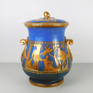 May include: A cobalt blue ceramic lidded urn featuring ornate gold-toned classical figures, trees, and architectural columns. The vessel includes two curved gold handles on the sides, a matching lid with a knob, and a decorative wave pattern border.