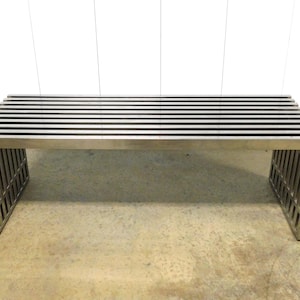 May include: A modern, stainless steel bench with a slatted top and geometric legs. The bench has a minimalist design and is suitable for indoor or outdoor use. The bench is approximately 60 inches long.