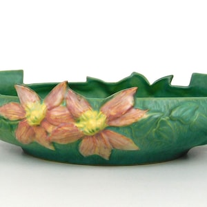 Pode incluir: Uma tigela de cerâmica verde com um design floral. A tigela apresenta duas grandes flores rosa e amarelas. A tigela tem uma borda ondulada e alças. A tigela está em uma superfície branca.