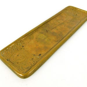 May include: A rectangular, antique brass tray with ornate, raised floral designs at each end. The tray has a slightly raised edge and a tarnished, aged appearance, suggesting it is vintage or an antique piece.