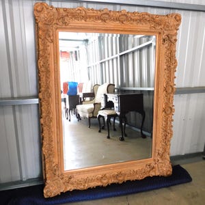 May include: Large, ornate, rectangular mirror with a detailed, light brown frame. The frame features intricate carvings and a decorative border. The mirror is resting on a blue moving blanket, reflecting furniture in the background.