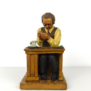 May include: A figurine of a jeweler at work, seated behind a wooden desk. The jeweler wears a yellow shirt, brown vest, and black pants. He is using a magnifying glass to examine a small object. The desk has a small tool and green objects on top.