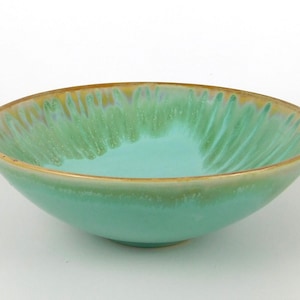 May include: A turquoise ceramic bowl with a gold rim. The bowl has a textured interior and a smooth exterior. The bowl is a light green color and is likely used for serving food.