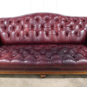 May include: A burgundy leather Chesterfield sofa with a button-tufted back and arms. The sofa has a wooden base and is set against a white wall. The sofa is a classic piece of furniture.