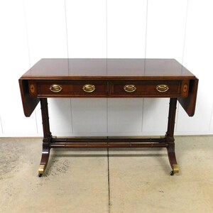 May include: A rectangular, dark brown wooden table with two drawers and brass hardware. The table has drop leaves on each side and stands on four legs with brass casters. The table is against a white wall.