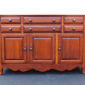 May include: A wooden sideboard featuring a warm brown finish, three upper drawers, two middle drawers, and three lower cabinet doors. The piece includes dark metal hardware, including round knobs on the drawers and decorative plates on the doors.