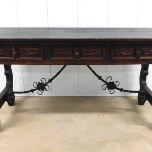 May include: A rectangular dark brown wooden console table featuring three drawers with carved front panels. The table is supported by four curved legs connected by two crossed black metal stretchers with decorative floral metal accents.