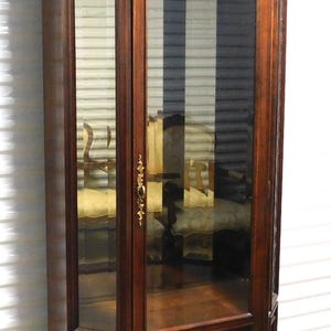 May include: A tall, dark wood corner cabinet with glass doors and ornate brass hardware. The cabinet features a decorative shell-shaped carving at the top and a paneled base. Interior shelves are visible through the glass.