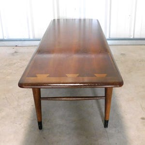 May include: A mid-century modern coffee table with a rectangular top and tapered legs. The table is made of wood with a dark brown finish and a decorative inlay. The legs are approximately 16 inches tall.
