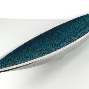 May include: An elongated, boat-shaped decorative bowl with a smooth, metallic silver exterior and a deep interior lined with a mosaic of small, multi-tonal blue and teal stone fragments set in a dark grout, creating a textured surface.