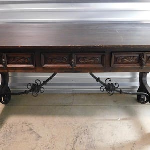 May include: Dark brown wooden desk with three drawers and ornate carvings. The desk has a rectangular top and is supported by carved legs and decorative metal supports. The desk is approximately 60 inches long and 30 inches wide.