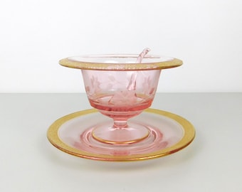 May include: A pink glass serving dish with a matching saucer and spoon. The dish and saucer have a gold rim. The dish has a pedestal base and a lid. The glass is etched with a floral design.