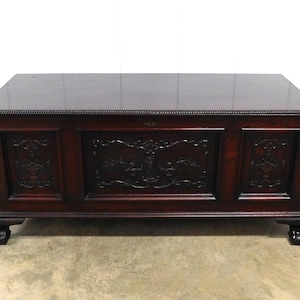 May include: A dark mahogany chest with a glossy top and ornate carved panels. The chest features detailed relief carvings and rests on four claw feet. A classic piece of furniture.