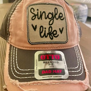 May include: A pink and gray distressed baseball cap with a white patch that says "Single life" in black script. The cap has a black label that says "OTTO Relaxed Fit OSFM Dad Hat Maximum Comfort Casual Style Pre-Washed Premium Fabric".