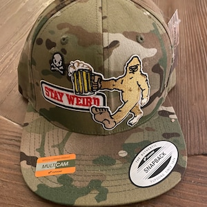 May include: Camouflage baseball cap with a patch featuring a Bigfoot character holding a beer mug, with the words "STAY WEIRD". The cap has a flat brim and a snapback closure. Includes a "MULTICAM" sticker.