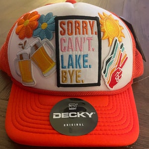 Puede incluir: Gorra de camionero naranja y blanca con parches bordados. Los parches incluyen flores, jarras de cerveza, un sol y un signo de la paz. La gorra presenta el texto "SORRY. CAN'T. LAKE. BYE."
