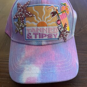 Puede incluir: Gorra de béisbol teñida con tinte anudado en tonos morado, rosa y azul. La gorra presenta un parche bordado con una puesta de sol, un cóctel y el texto "TANNED & TIPSY". Adornada con estrellas de mar de pedrería.