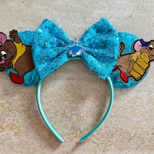 Könnte beinhalten: Ein hellblaues Stirnband mit Pailletten-Schleife und gestickten Cartoon-Mäusen auf jeder Seite. Die Schleife hat einen kleinen Cinderella-Anhänger. Ein lustiges Accessoire für Disney-Fans.