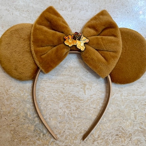 May include: Brown plush mouse ear headband with a large bow and a Simba and Nala pin. The ears and bow are a solid brown colour, and the headband is a light tan. The pin features the characters from the Lion King.