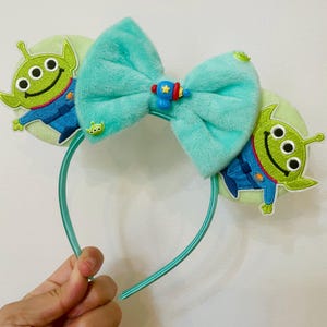 May include: A teal headband featuring plush ears with embroidered green aliens in blue uniforms. A large teal bow with a small blue and red spaceship accent is centered on the headband. The headband is held by a hand.