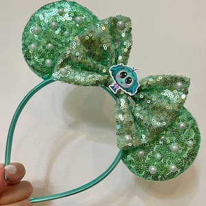 May include: Green sequined Minnie Mouse ears headband with a green bow and a small character charm. The bow has white pearl accents.