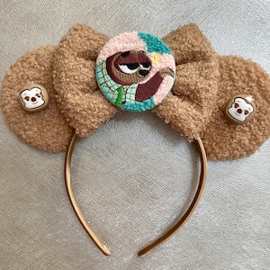 May include: Brown plush headband with large bow-shaped ears. The center features a round patch with an embroidered sloth character. Small bear-shaped buttons adorn each ear. The headband is gold-colored.