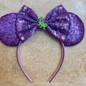 Peut inclure: Serre-tête oreilles de souris à paillettes violettes avec un grand nœud et un charm caméléon vert. Les oreilles et le nœud sont recouverts de paillettes scintillantes. De petites fleurs blanches ornent le nœud et les oreilles. Le bandeau est violet clair.