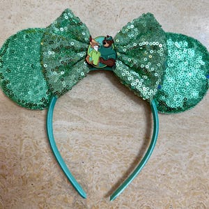 May include: A teal sequined headband with large ears and a bow. The bow features a circular emblem with cartoon characters. The ears and bow are covered in teal sequins. The headband is teal.