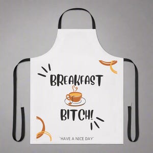 May include: White apron with black straps featuring the text "BREAKFAST BITCH!" in black, with a brown coffee cup graphic. The phrase "HAVE A NICE DAY" is printed at the bottom. Decorative orange and brown accents surround the text.