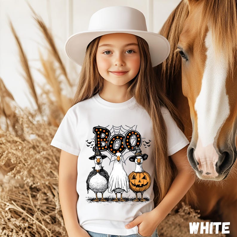 K&ouml;nnte beinhalten: Ein Kind tr&auml;gt ein wei&szlig;es T-Shirt mit einem Halloween-Motiv. Das Shirt zeigt das Wort "Boo" &uuml;ber drei Cartoon-G&auml;nsen, die als Hexe, Geist und K&uuml;rbis verkleidet sind. Das Kind tr&auml;gt auch einen wei&szlig;en Hut.