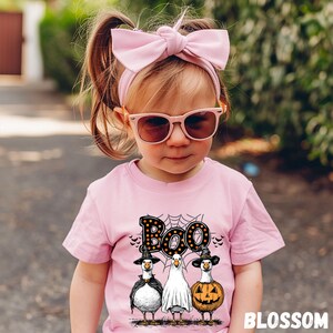 K&ouml;nnte beinhalten: Ein Kind tr&auml;gt ein rosa T-Shirt mit einem Halloween-Motiv. Das Shirt zeigt das Wort "Boo" &uuml;ber drei Cartoon-G&auml;nsen, die als Hexe, Geist und K&uuml;rbis verkleidet sind. Das Kind tr&auml;gt auch ein rosa Stirnband mit Schleife und Sonnenbrille.