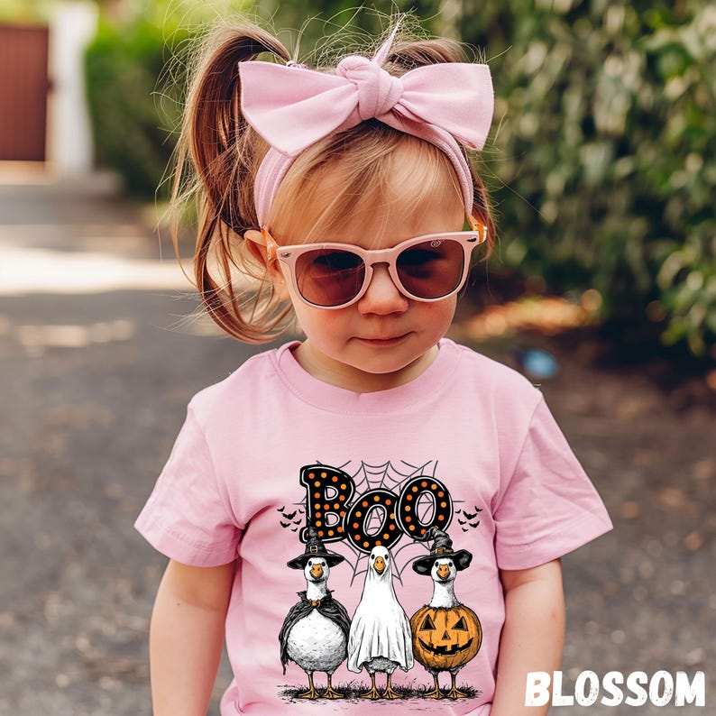K&ouml;nnte beinhalten: Ein Kind tr&auml;gt ein rosa T-Shirt mit einem Halloween-Motiv. Das Shirt zeigt das Wort "Boo" &uuml;ber drei Cartoon-G&auml;nsen, die als Hexe, Geist und K&uuml;rbis verkleidet sind. Das Kind tr&auml;gt auch ein rosa Stirnband mit Schleife und Sonnenbrille.