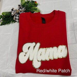 Könnte beinhalten: Ein rotes Sweatshirt mit dem Wort "Mama" in weißen, flauschigen Buchstaben, die goldfarben umrandet sind. Der Text "Red/white Patch" befindet sich unten. Das Sweatshirt ist auf einer weißen Oberfläche gefaltet, mit einer dekorativen Schleife und Grünzeug im Hintergrund.