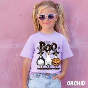 K&ouml;nnte beinhalten: Ein helllila T-Shirt mit einem Halloween-Design. Die Grafik zeigt das Wort "Boo", ein Spinnennetz und drei Cartoon-G&auml;nse in Kost&uuml;men: eine Hexe, ein Geist und ein K&uuml;rbis. Das M&auml;dchen tr&auml;gt eine Sonnenbrille. Das Wort "ORCHID" steht unten.