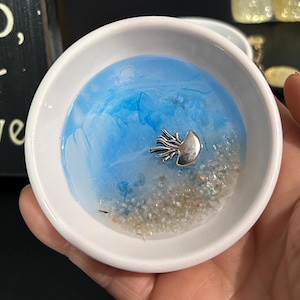 Op de afbeelding: Een kleine, witte keramische schaal met een blauw en wit harsinterieur, dat op zeewater lijkt. Een zilverkleurige kwalbedel rust op een bedje van zandachtige korrels. De schaal wordt in een hand gehouden.