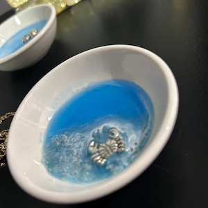 Può includere: Due piccole ciotole ovali in ceramica bianca con un design in resina blu e bianco. Ogni ciotola presenta un ciondolo a forma di granchio color argento. Le ciotole sono su una superficie nera, con una catena d'oro visibile.