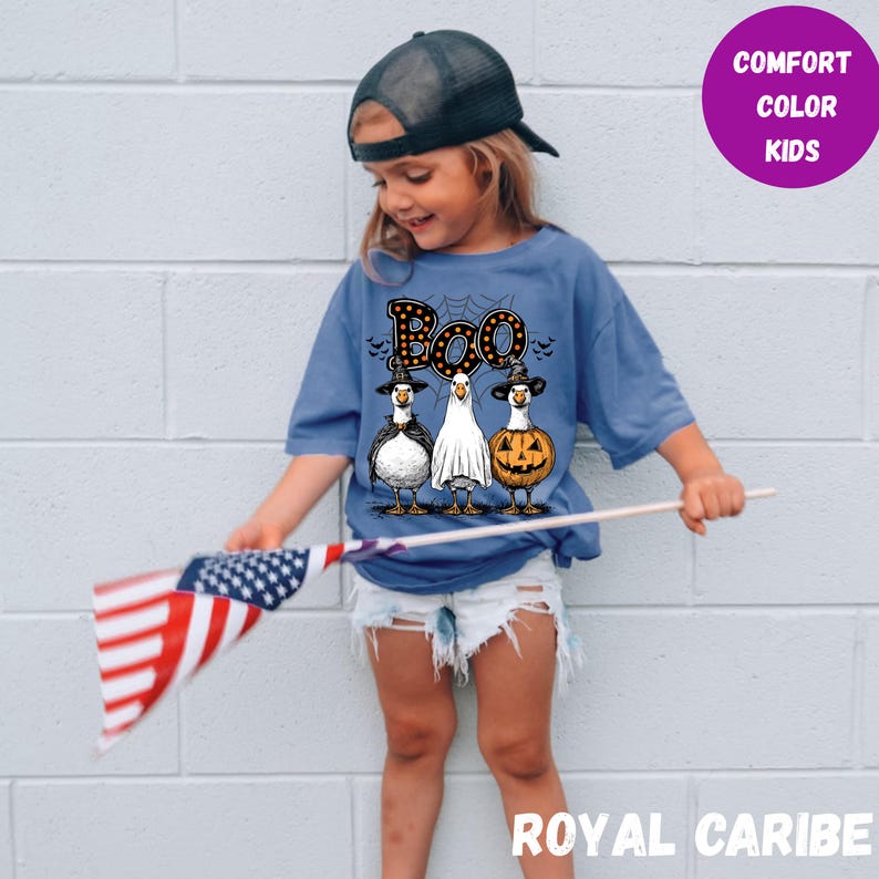 K&ouml;nnte beinhalten: Ein blaues Comfort Color Kinder-T-Shirt mit Halloween-Motiv. Das Shirt zeigt drei Cartoon-V&ouml;gel, die als Vampir, Geist und K&uuml;rbis verkleidet sind, mit dem Wort "Boo" dar&uuml;ber. Das Kind h&auml;lt eine amerikanische Flagge.