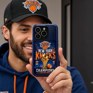 May include: A dark blue smartphone case featuring a New York Knicks graphic with a gold championship trophy, basketball imagery, and the text New York Knicks Champions. The case has a triple camera cutout and a raised border around the lens array.
