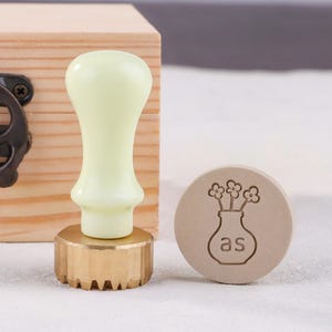 May include: A light green wax seal stamp with a brass base and a wooden box in the background. The stamp has a floral design with the letters "as" inside a vase. The stamp is for crafts and stationery.