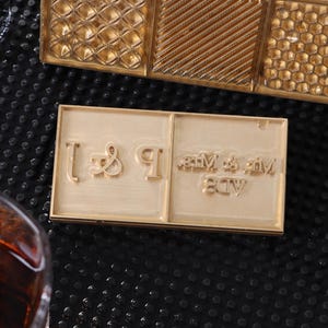 Puede incluir: Primer plano de un sello dorado con el texto "Mr & Mrs" y otros elementos decorativos. El sello es rectangular y tiene un diseño en relieve. El fondo es una superficie oscura y texturizada.