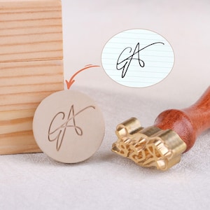 Puede incluir: Un sello de cera personalizado con un mango de madera y un cabezal de sello de latón. El sello presenta las iniciales "GA" en una escritura elegante. Una caja de madera está en el fondo. El diseño también se muestra en un pequeño círculo de papel rayado.