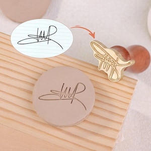 May include: A custom signature stamp with a wooden handle and a circular stamp head. The stamp is made of brass and features a unique signature design. The stamp is shown next to an impression of the signature on a light brown surface.
