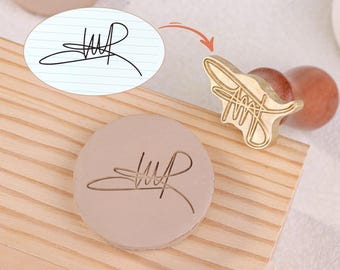 以下が含まれることがあります： 木製のハンドルと円形のスタンプヘッドが付いた、カスタム署名スタンプ。スタンプは真鍮製で、ユニークな署名デザインが特徴です。スタンプは、明るい茶色の表面に署名の印象の隣に表示されます。