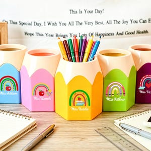 May include: Five pencil-shaped desk organizers in light blue, pink, yellow, green, and purple. Each features a rainbow and apple graphic with a name printed below. The yellow organizer holds a collection of colored pencils. The items are arranged in a row.