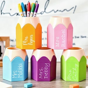 Puede incluir: Cinco organizadores de escritorio con forma de lápiz en naranja, rosa, azul claro, morado y verde lima. Cada recipiente tiene una parte superior color melocotón claro y está personalizado con texto blanco: Mrs. Agnes, Mrs. Rachel, Mrs. Matilda, Mrs. Debby y Mrs. Cathy.