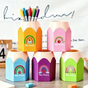 May include: Five hexagonal pencil-shaped pen holders in yellow, pink, blue, purple, and green. Each holder features a rainbow and apple graphic with a name printed below. The yellow holder contains five colored markers. The design mimics a pencil tip.