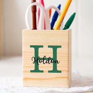 Puede incluir: Un portalápices de madera con la letra "H" y el nombre "Holdon" en verde. El portalápices contiene bolígrafos, lápices y tijeras. Una mano sostiene un bolígrafo naranja. El portalápices está sobre una superficie de encaje blanco.